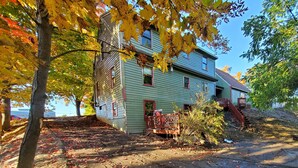 A two-story house with a green exterior and red trim, surrounded by trees with autumn leaves. - Serenity between the lakes and seashore (Westbrook)