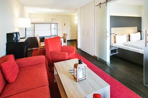 Et moderne hotelværelse med en rød sofa, et træbord, en seng med hvide sengetæpper og et tekøkken.