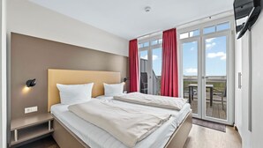 Habitación de hotel moderna con una cama grande, una mesita de noche, un televisor montado en la pared y un balcón con cortinas rojas.