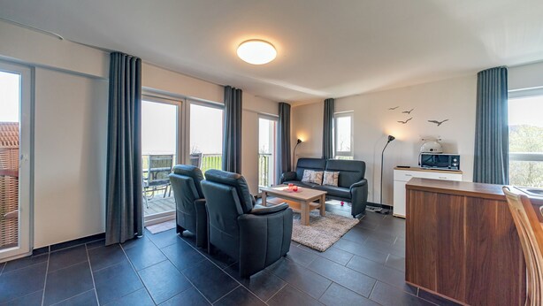 A modern living room with a sofa, two armchairs, a coffee table, and a wooden cabinet. There is a balcony with a view of the outside. - Exklusives Ferienapartment mit Meerblick, Balkon, Sauna und Whirlbadewanne (Nordseeheilbad Schillig)