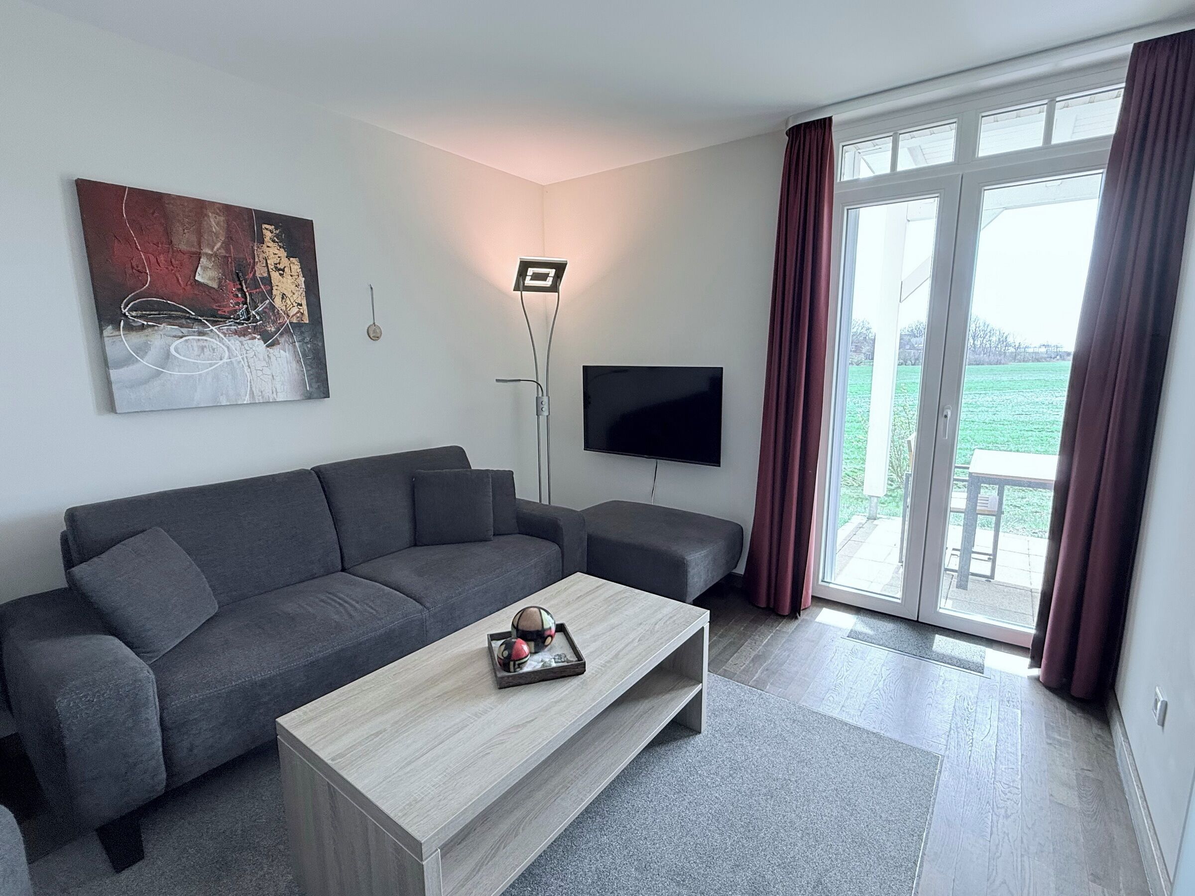 A modern living room with a grey sofa, a coffee table, a flat-screen TV, and a large window with curtains.