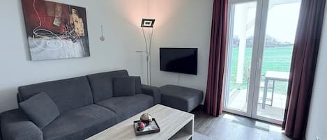 Ein modernes Wohnzimmer mit grauem Sofa, Couchtisch, Flachbildfernseher und einem großen Fenster mit Vorhängen.