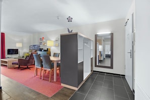 Una habitación de hotel moderna con zona de comedor, televisor, sofá y un espejo grande.