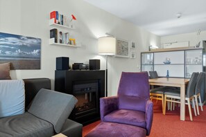 Una sala de estar moderna con chimenea, un sillón morado, un sofá, una mesa de comedor con sillas y una estantería con libros y objetos decorativos.