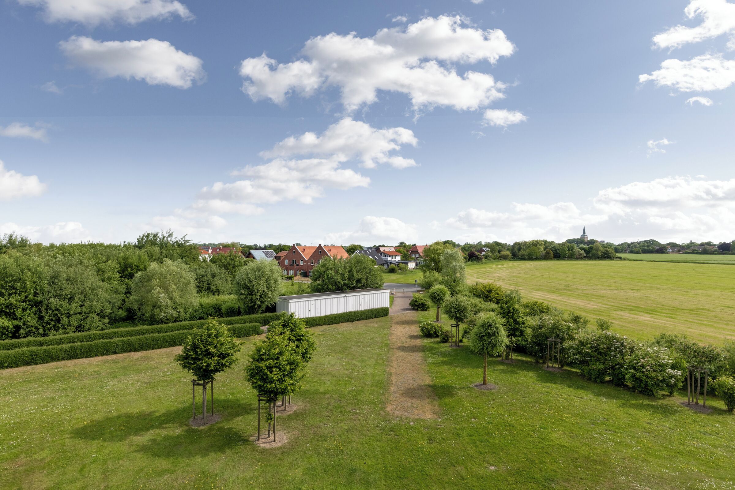 Un vaste champ herbeux et dégagé, avec un sentier menant à un bâtiment, le tout entouré d’arbres et sous un ciel clair.