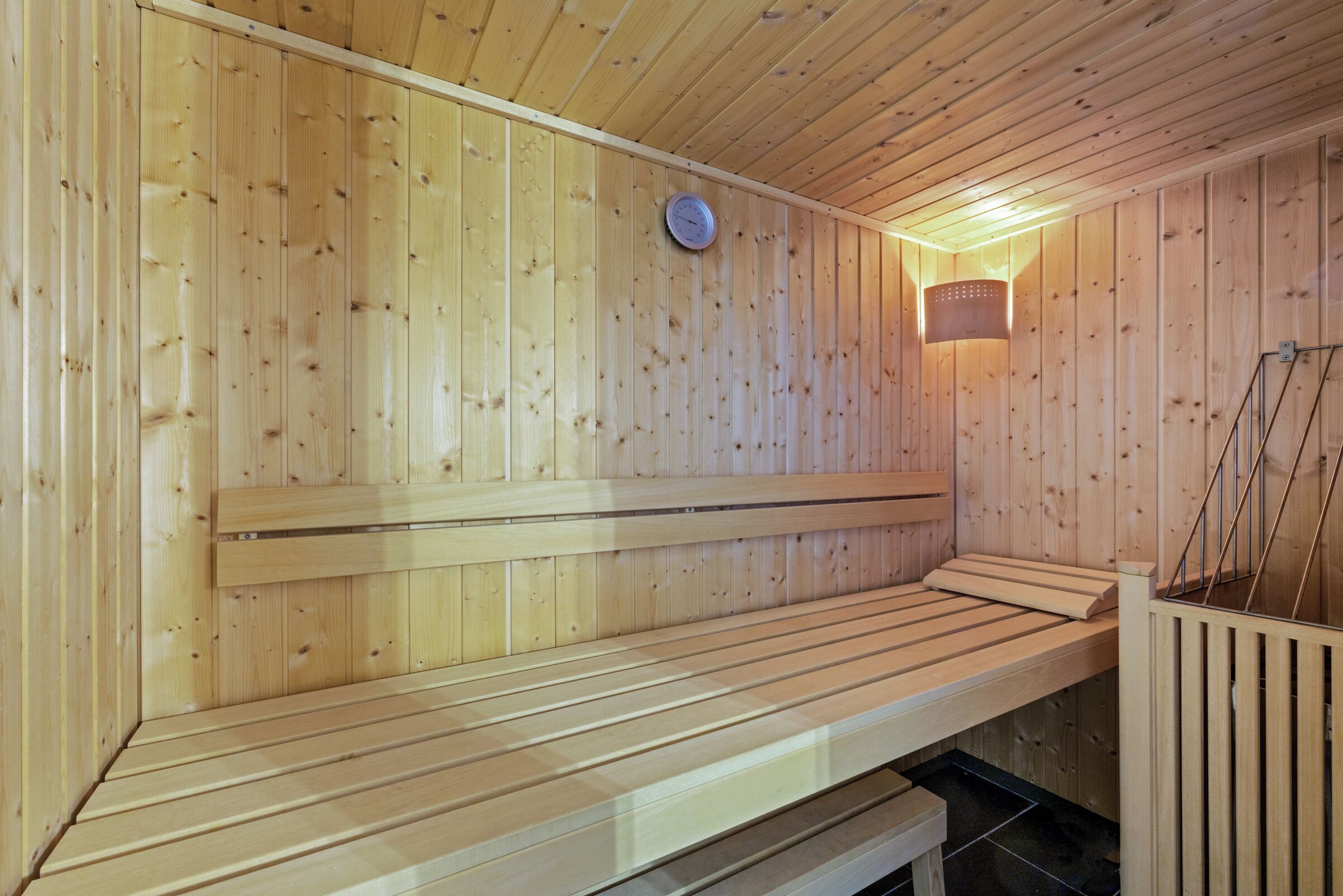 A sauna with wooden walls and benches, a clock, and a wall-mounted light fixture.
