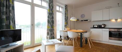 A modern kitchen with white cabinets, a dining area with a wooden table and chairs, a television on a stand, and large windows with curtains.