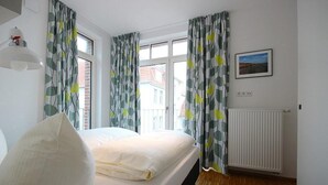 Un dormitorio con cama, cortinas, radiador y ventana con vista a edificios.