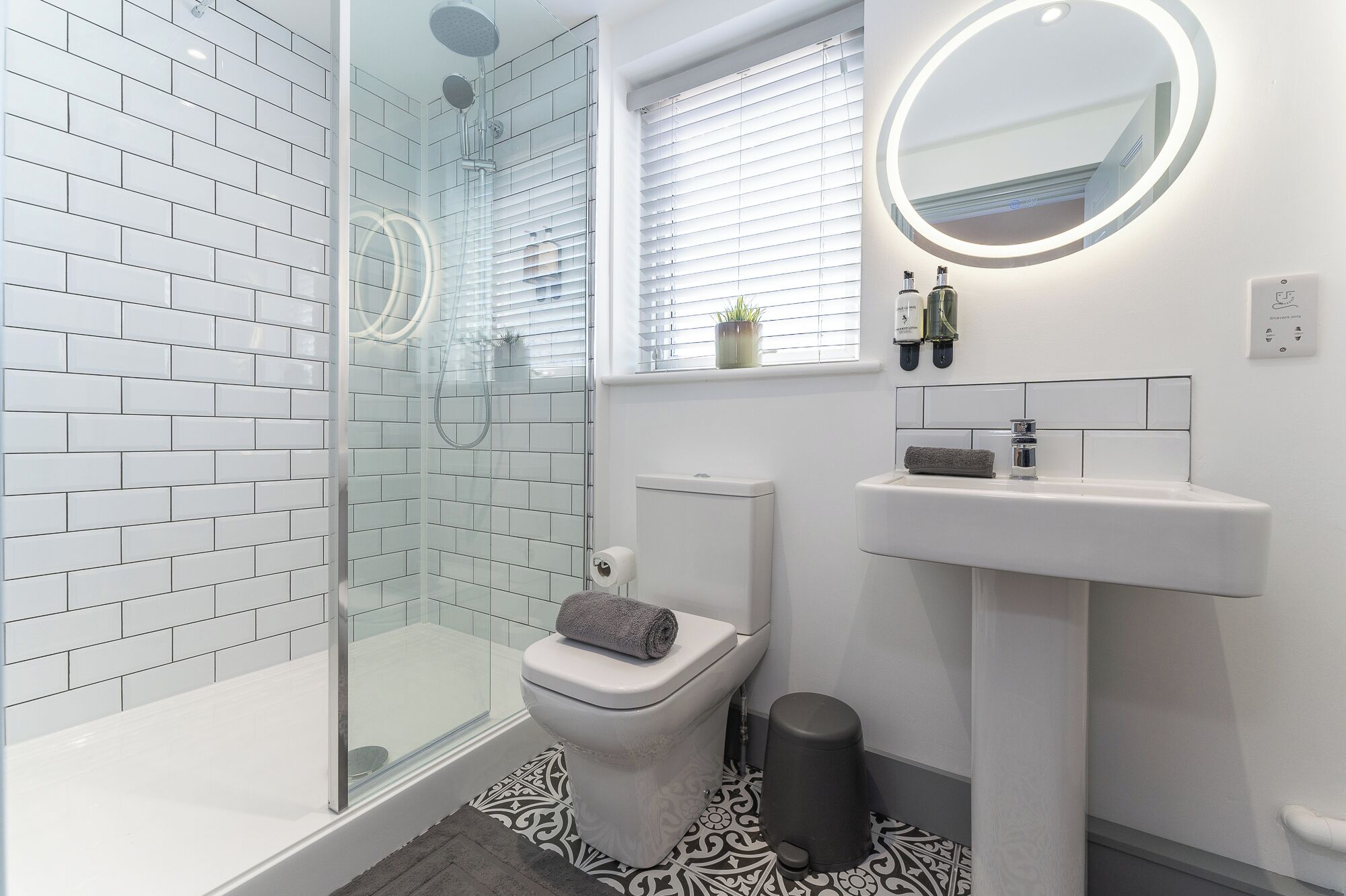 Un bagno moderno con doccia vetrata, pareti rivestite di piastrelle bianche, un lavandino bianco e uno specchio rotondo con cornice illuminata.