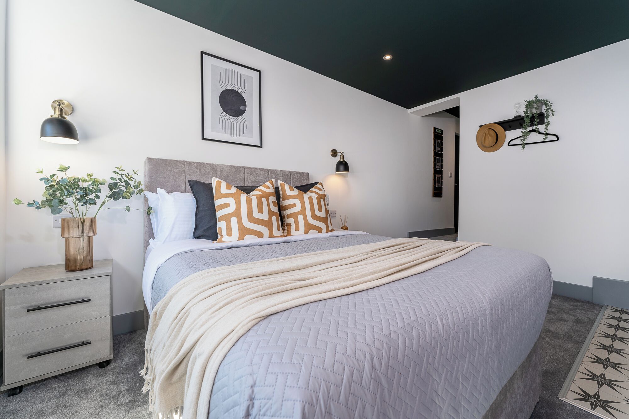 A modern bedroom with a bed, bedside tables, a wall-mounted lamp, and a framed artwork.