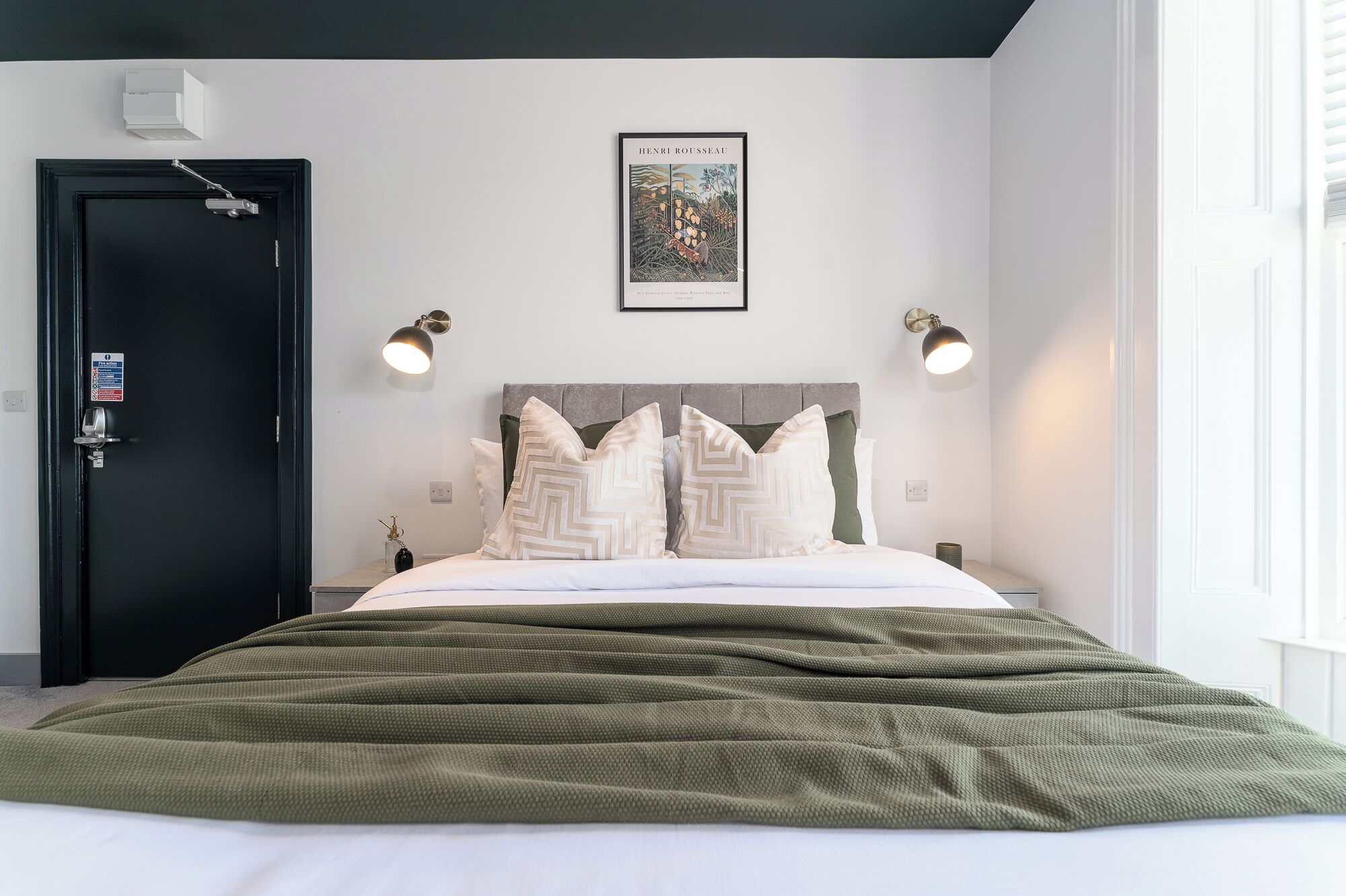 A bedroom with a bed, a black door, a framed picture on the wall, and two wall-mounted lamps.