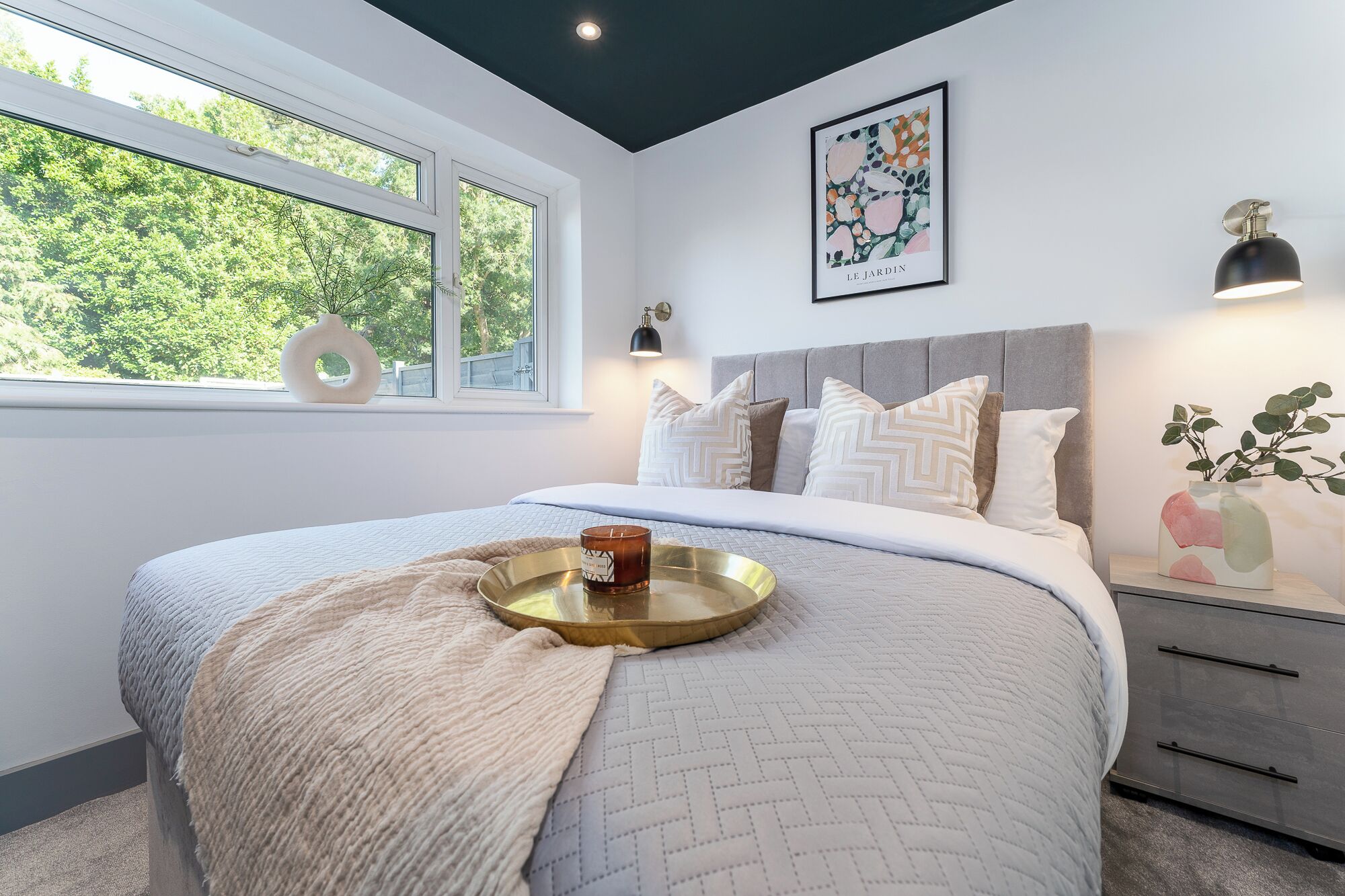 Quarto moderno com uma cama grande, uma mesa de cabeceira com abajur, uma janela com vista para o verde e uma obra de arte emoldurada na parede.