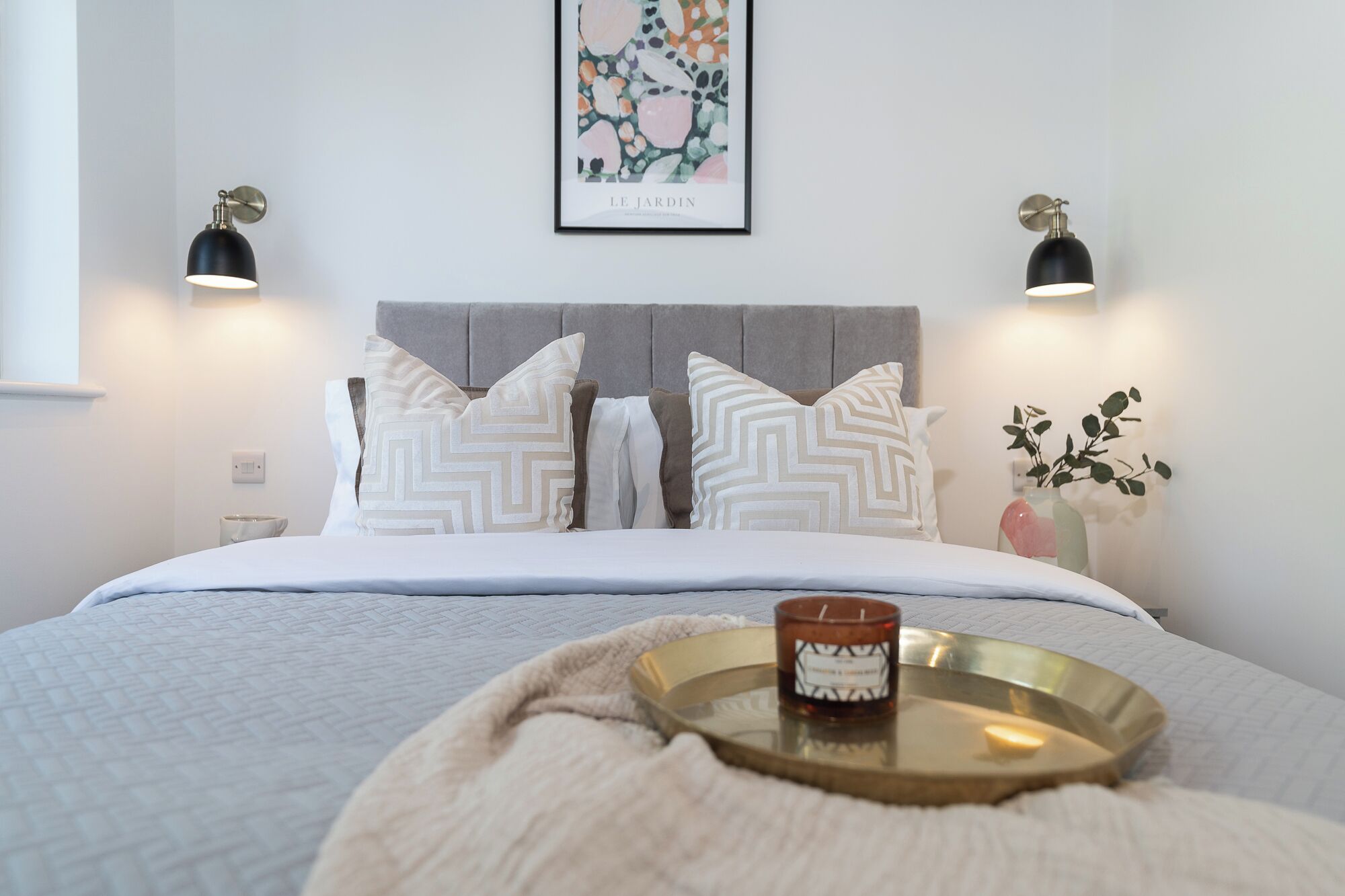 Un letto rifatto con cuscini decorati, un quadro appeso sopra e due lampade fissate al muro.