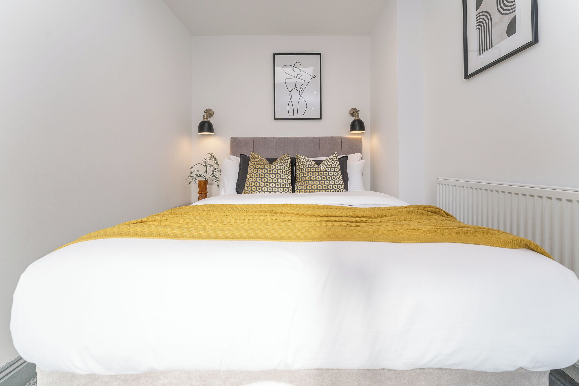 A neatly made bed with a white and mustard blanket, a grey headboard, and patterned pillows. There are two wall-mounted lamps and a framed abstract artwork above the bed.