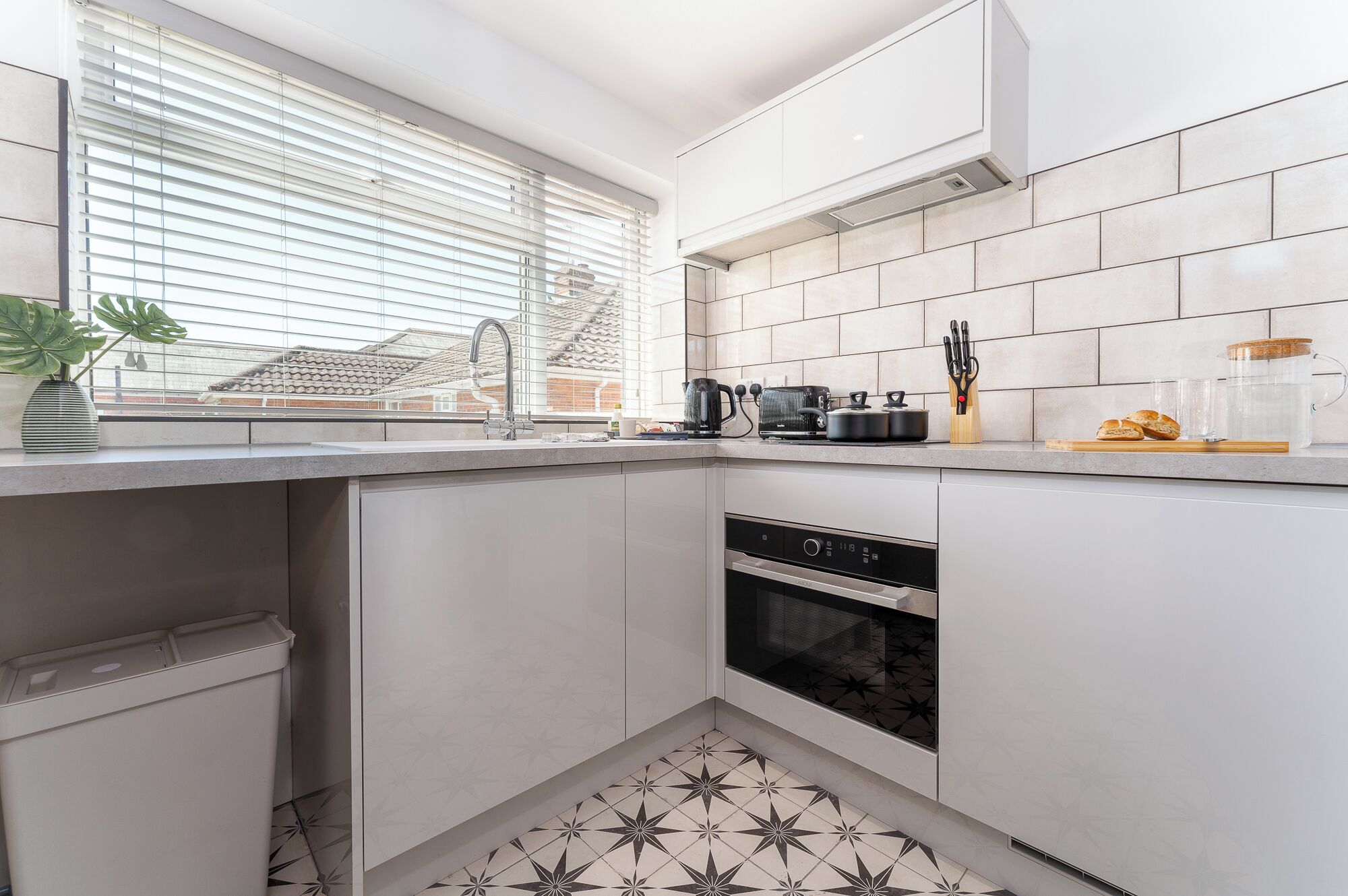 A modern kitchen with white cabinets, a built-in oven, a coffee maker, and a window with blinds.