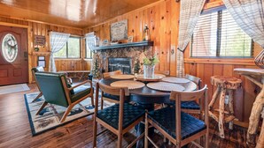 Interior de una cabaña de madera con una mesa redonda para comer, sillas tapizadas de verde, una chimenea y un árbol de Navidad.