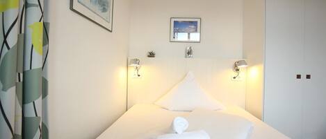 Habitación de hotel con cama, dos lámparas de pared, un pequeño cuadro enmarcado sobre la cama y una puerta a la derecha.
