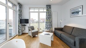 A modern living room with a sofa, armchair, and coffee table, large windows with curtains, a flat-screen TV, and a framed picture on the wall. - Tolles Ferienapartment mit Großem, Sonnigen Südbalkon - nur 100 m zum Strand! (Nordseeheilbad Wangerooge)
