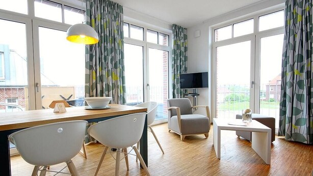 A modern living room with a wooden floor, a dining area with two white chairs, a grey sofa, a small white table, and a TV on a stand. - Tolles Ferienapartment mit Großem, Sonnigen Südbalkon - nur 100 m zum Strand! (Nordseeheilbad Wangerooge)