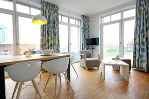 Uma sala de estar moderna com piso de madeira, uma área de jantar com duas cadeiras brancas, um sofá cinza, uma pequena mesa branca e uma TV sobre um suporte.