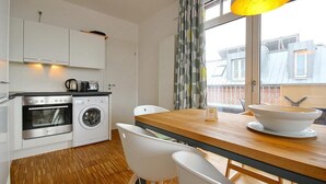 A modern kitchen with white cabinets, a wooden dining table, and a view of the city through large windows. - Tolles Ferienapartment mit Großem, Sonnigen Südbalkon - nur 100 m zum Strand! (Nordseeheilbad Wangerooge)