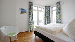 A bedroom with a bed, a chair, and a window with curtains. - Tolles Ferienapartment mit Großem, Sonnigen Südbalkon - nur 100 m zum Strand! (Nordseeheilbad Wangerooge)