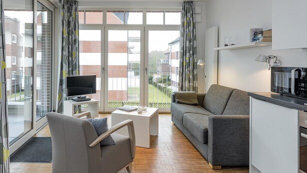 A modern apartment with a grey sofa, a small white table, a TV, and a kitchen area.