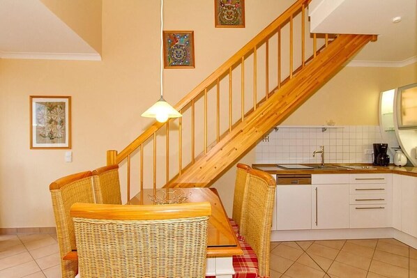 Una cocina con una escalera de madera, sillas de mimbre, una mesa de comedor y una lámpara colgante.