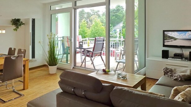 Una sala de estar moderna con un sofá, una mesa de comedor con sillas, un televisor y ventanas amplias que dan al exterior.