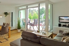 Un salon moderne comprenant un canapé, une table à manger avec des chaises, un téléviseur et de grandes fenêtres donnant sur l’extérieur.