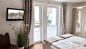 Un dormitorio con una cama, un televisor en la pared, una planta en maceta y una ventana con cortinas.