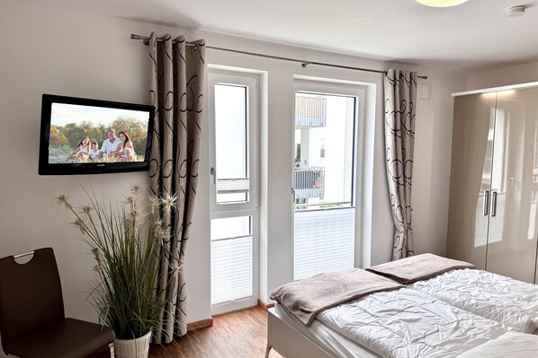 Une chambre à coucher avec un lit, un téléviseur fixé au mur, une plante en pot et une fenêtre avec des rideaux.