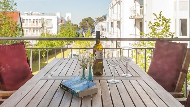 A wooden table with a wine bottle, two glasses, and a book on a balcony overlooking a cityscape.