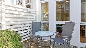 Un área de estar al aire libre con una mesa redonda y dos sillas, un cerco blanco y un ventanal que da a una habitación con cama y ventana.