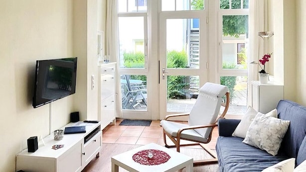 Un salón con televisor, muebles blancos, un sofá azul y vistas al exterior a través de amplias ventanas.