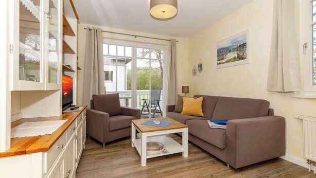 A cozy living room with a sofa, armchair, and a wooden coffee table. There is a window with curtains, a radiator, and a picture on the wall.