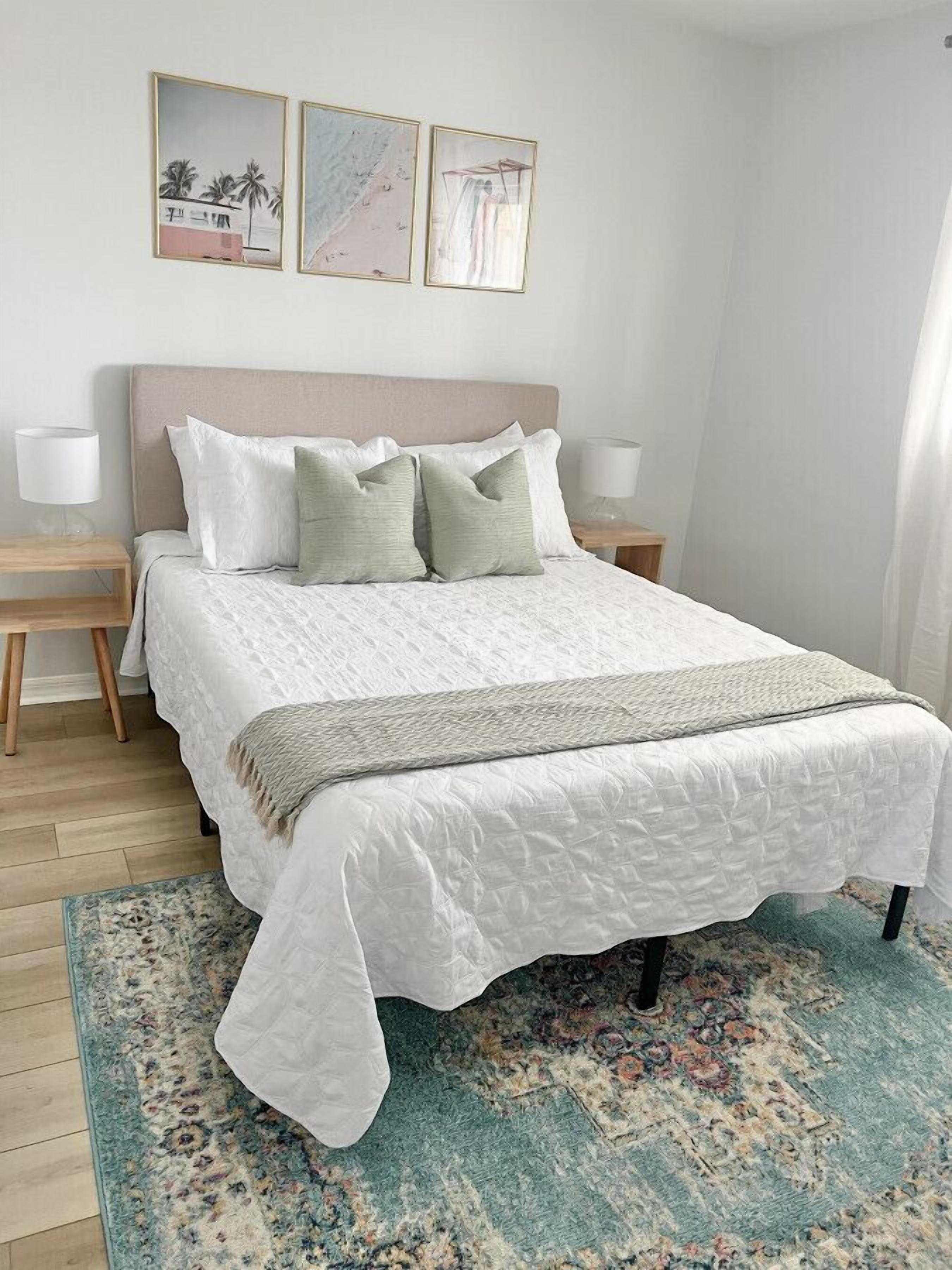 A neatly made bed with a white duvet and green pillows, a patterned rug, and framed artwork on the wall.