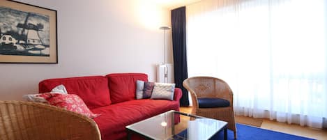 Una sala de estar con un sofá rojo, dos sillones de mimbre, una mesa de centro de vidrio y un cuadro en la pared.