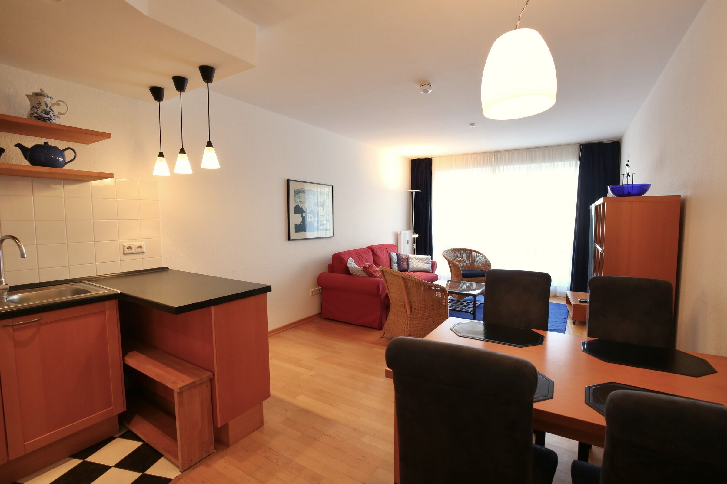 A compact kitchen with a sink, wooden cabinets, and a checkered floor. A dining area with a table and chairs is visible, along with a red sofa and a wicker chair.