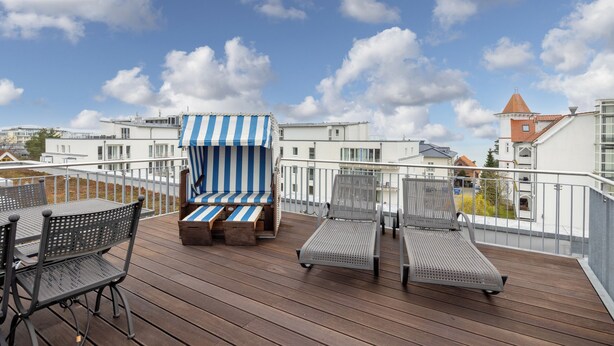 A wooden deck with lounge chairs and a striped beach chair, overlooking a cityscape with buildings and a clear sky.