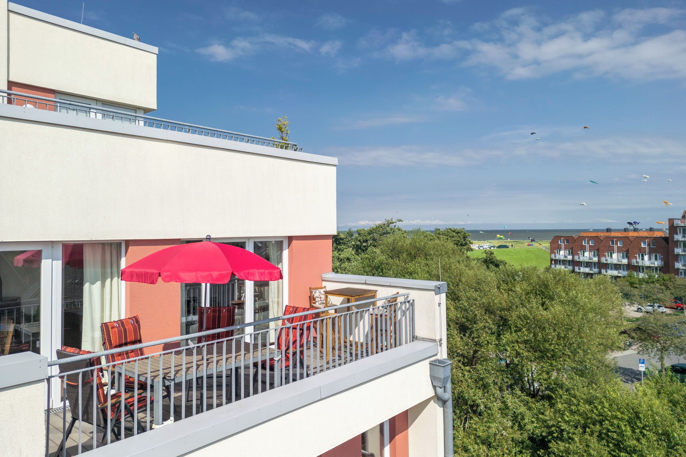A balcony with outdoor furniture and a red umbrella, overlooking a residential area with trees and buildings.