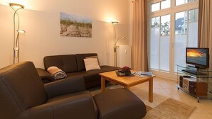 Una sala moderna con sofá, mesa de centro y televisor. Hay un cuadro en la pared y una ventana con cortinas.