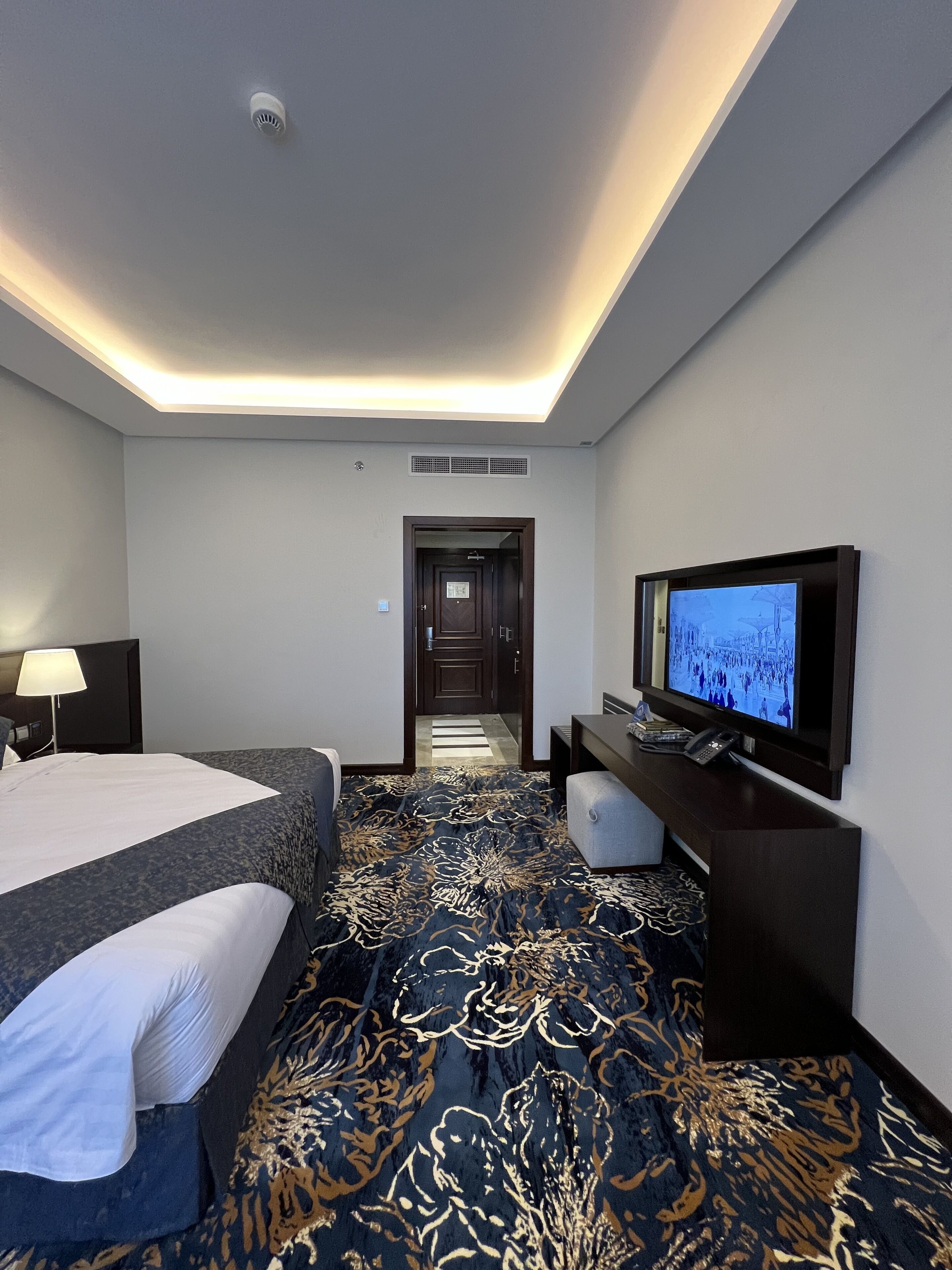 Quarto de hotel com cama, escrivaninha com televisão, candeeiro e tapete estampado.