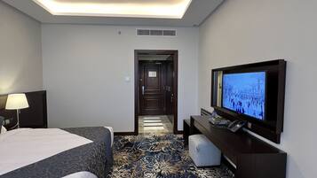 Quarto de hotel com cama, escrivaninha com televisão, candeeiro e tapete estampado.