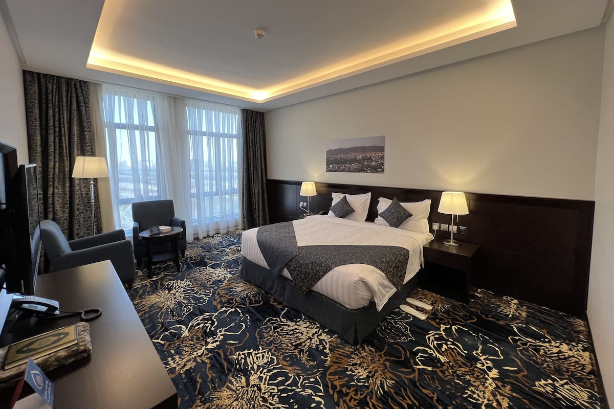 Habitación de hotel con una cama grande, dos mesitas de noche con lámparas, un escritorio con silla, un televisor y vista a un paisaje urbano a través de la ventana.