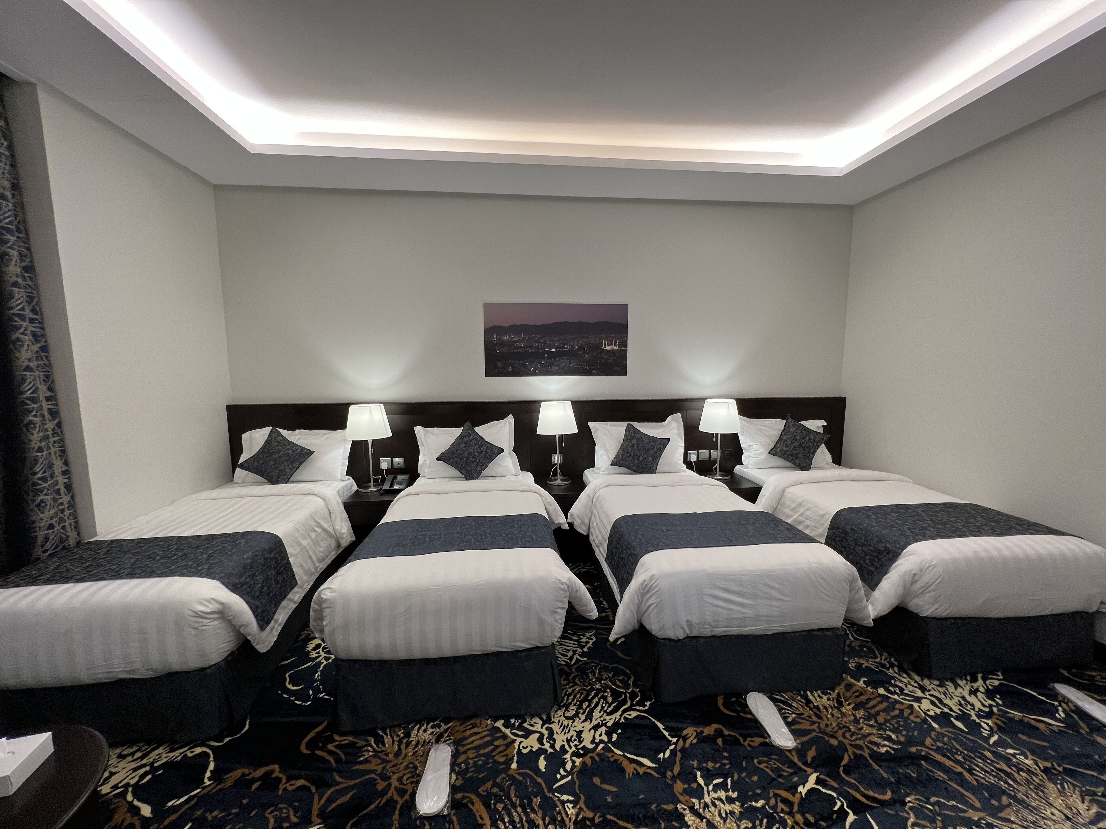 Habitación de hotel con cuatro camas dispuestas en fila, vista panorámica de la ciudad y alfombra estampada.