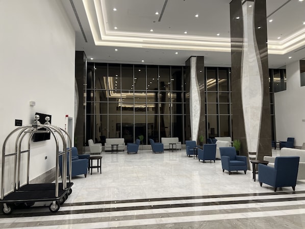 A spacious lobby with a modern design, featuring a seating area with blue and white chairs, a reception desk, and a large glass wall providing a view of the interior.