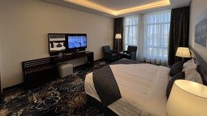 A hotel room with a large bed, a desk with a flat-screen TV, two armchairs, a small table, and a window with curtains. - Zaha Taiba Hotel (Madinah)