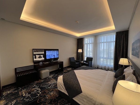 A hotel room with a large bed, a desk with a flat-screen TV, two armchairs, a small table, and a window with curtains. - Zaha Taiba Hotel (Madinah)