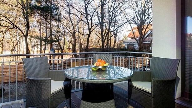 A round glass table with wicker chairs on a wooden deck, surrounded by a metal railing. - Modernes, Strandnahes Apartment mit Großem Balkon, Fußbodenheizung u. Kaminofen (Ostseebad Binz)